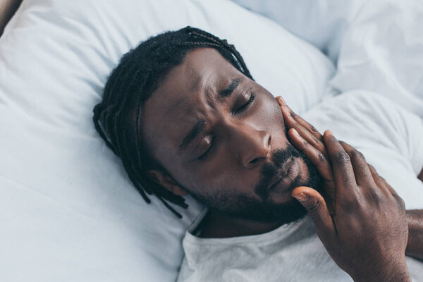 unhappy african american man suffering from jaw pain while lying in bed