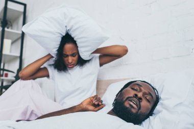 unhappy african american woman covering head with pillow while sitting in bed near snoring husband clipart