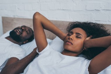 exhausted african american woman covering ears with hands while lying near snoring husband  clipart