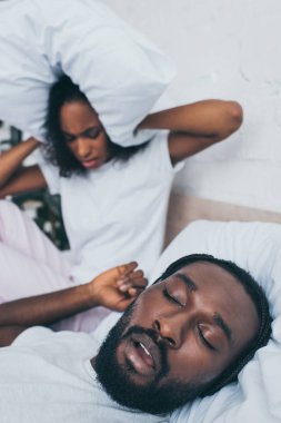 selective focus of african american woman covering head with pillow while sitting in bed near snoring husband clipart