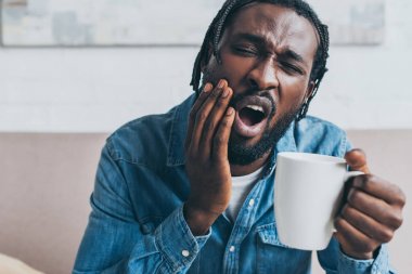 Genç Afrikalı Amerikalı bir adam diş ağrısı çekerken elinde kahve fincanı tutuyor.