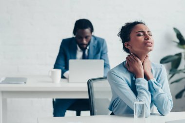 selective focus of african american businesswoman suffering from neck pain while sitting near manager working at laptop  clipart