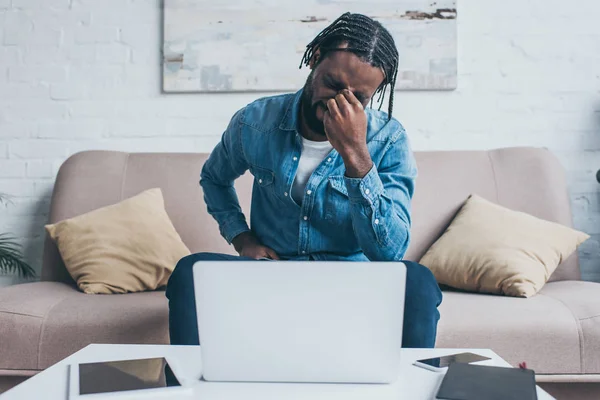 デジタルデバイスでテーブルの近くに座っている間 頭痛に苦しむ疲れたアフリカ系アメリカ人男性 — ストック写真