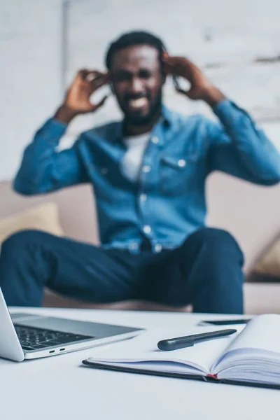 ノートパソコンとノートパソコンでテーブルの近くに座っている間 頭痛に苦しむアフリカ系アメリカ人男性の選択的な焦点 — ストック写真