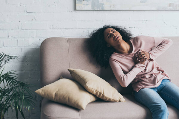 unhappy african american woman suffering from heart pain while sitting on sofa 