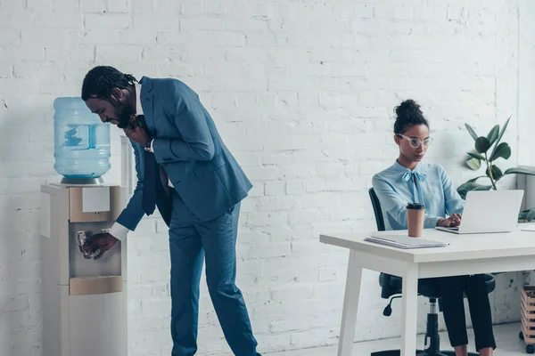 Thirsty African American Manager Standing Water Cooler Colleague Sitting Workplace — Stock Photo, Image