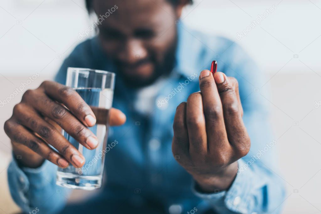 selective focus of african american man suffering from hurt and holding glass of water and pill