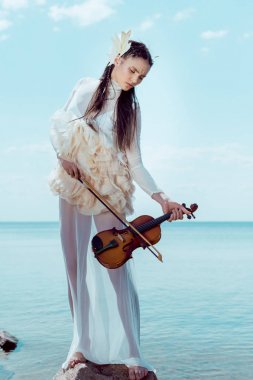 tender woman in white swan costume with violin standing on blue river and sky background clipart