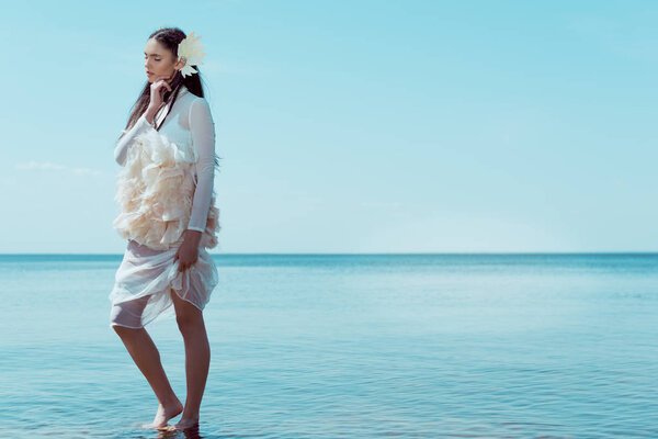 brunette woman in white swan costume closing eyes, standing on river and sky background 