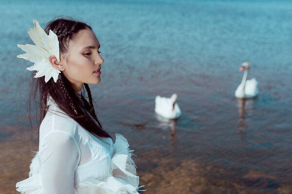 side view of tender woman in white swan costume looking away, standing on background with birds and river