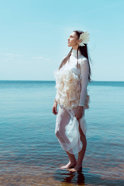 young woman in white swan costume standing on river and sky background, closing eyes