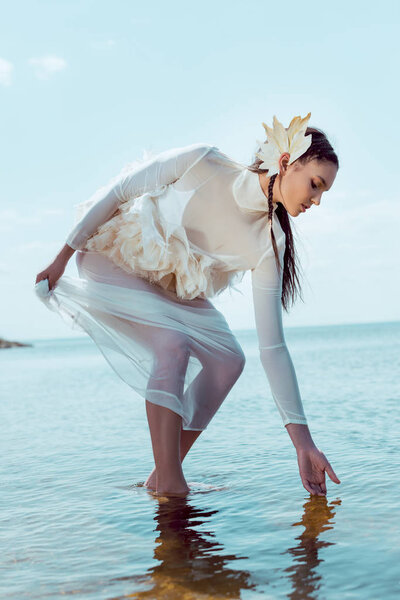 elegant woman in white swan costume touching river, looking at water
