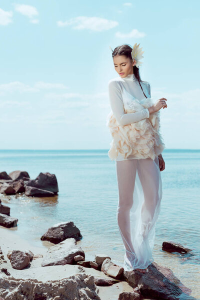 tender woman in white swan costume looking away, standing on beach near water