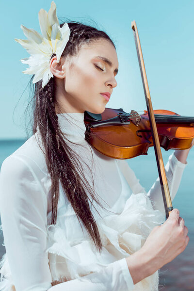 portrait of beautiful woman in white swan costume with violin closing eyes, playing music on sky background