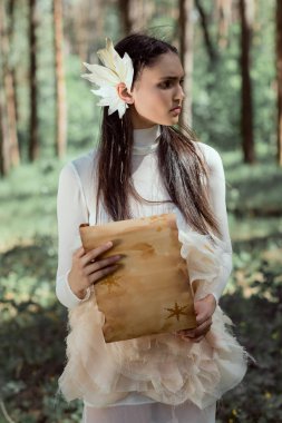 orman arka plan üzerinde duran parşömen ile beyaz kuğu kostümü zarif kadın, uzağa bakıyor