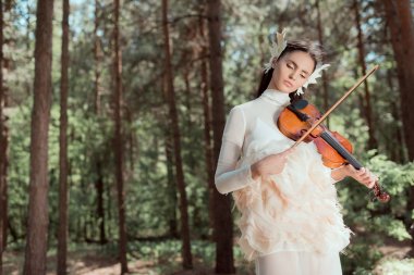 dreamy woman in white swan costume standing on forest background, playing on violin clipart