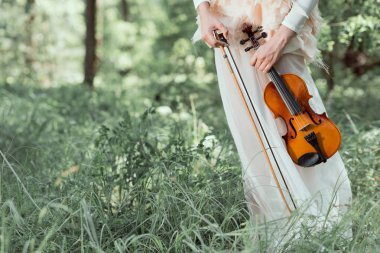 cropped view of woman in white swan costume standing on forest background, holding violin clipart