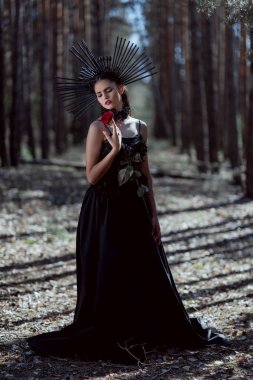 young woman in witch costume closing eyes, holding red rose, standing on forest background clipart