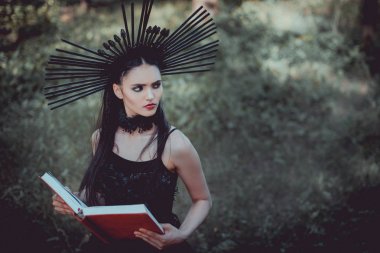 orman arka planda duran cadı kostümlü güzel kadın yüksek açı görünümü, kırmızı kitap tutarak, uzağa bakarak