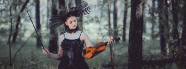 panoramic view of woman in witch costume with crown standing on forest background, holding violin clipart