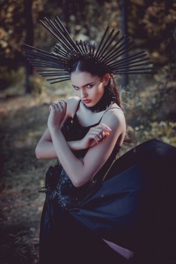 Siyah cadı kostümlü genç bir kadın orman arka planında kameraya bakıyor.