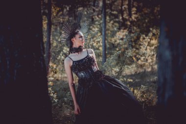 selective focus of beautiful woman in black witch costume standing between trees clipart