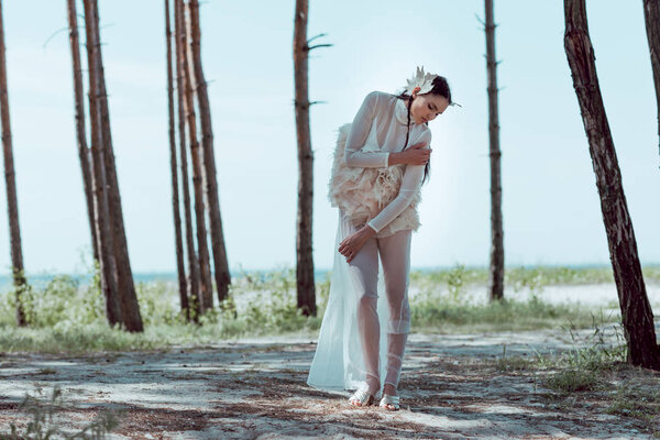 beautiful woman in white swan costume standing on sandy beach near trees