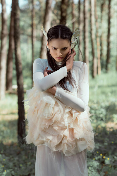 adult woman in white swan costume standing on forest background, holding hands near face