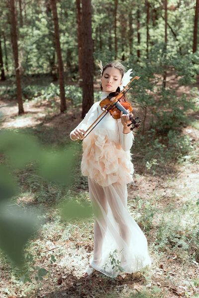selective focus of brunette woman in white swan costume standing on forest background, playing on violin