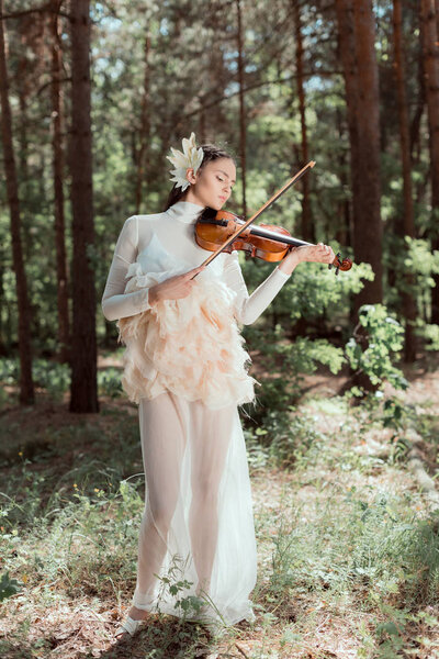 elegant woman in white swan costume standing on forest background, playing on violin