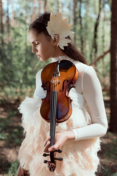 tender woman in white swan costume standing on forest background with violin, looking away