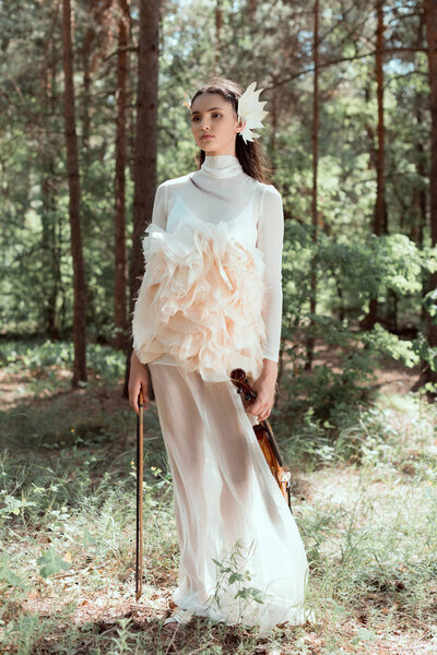young woman in white swan costume standing on forest background with violin, looking away