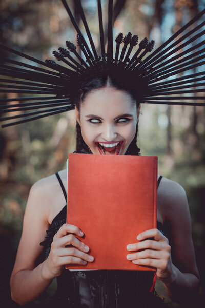 сумасшедшая женщина в черном костюме ведьмы с короной на голове гримаса, держа красную книгу
