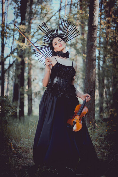 low angle view of young woman in witch costume standing on forest background, holding violin