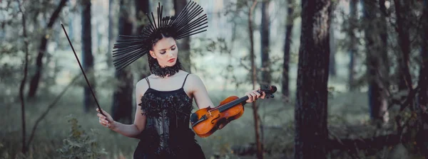 森の背景に立つ王冠を持つ魔女の衣装を着た女性のパノラマビュー バイオリンを保持 — ストック写真