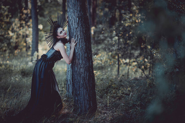 elegant woman in witch costume standing near tree trunk, looking at camera