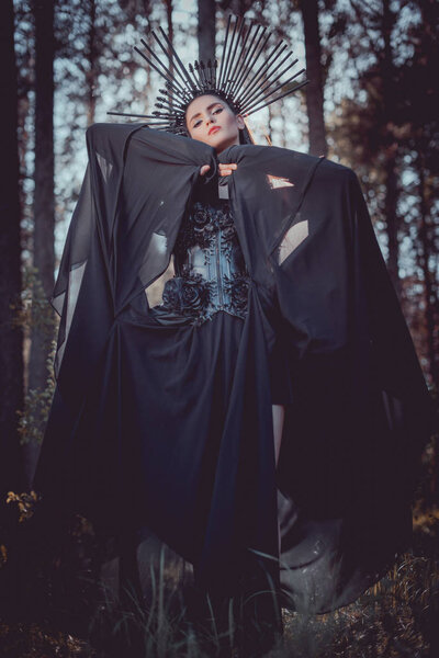 low angle view of woman in witch costume and crown on head holding hands with black textile near face
