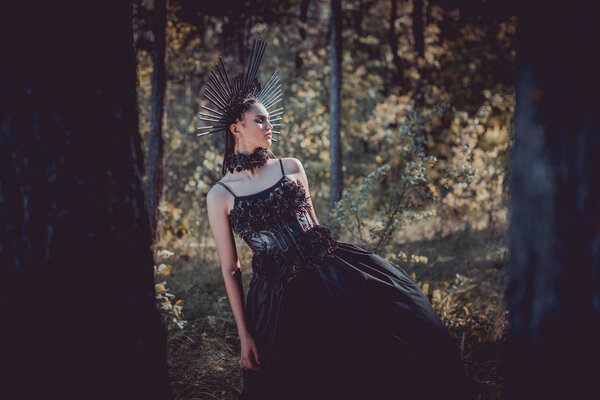 selective focus of beautiful woman in black witch costume standing between trees