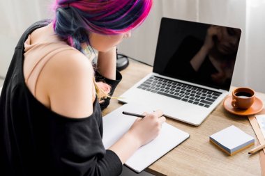 Back view of girl writing notes while sitting at table with laptop, sticky notes and cup of coffee clipart