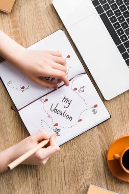 cropped view of girl drawing in notebook with my vacation lettering on wooden table clipart
