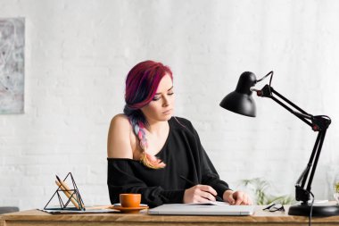 attractive hipster girl writing something while sitting behind table with laptop, cup of coffee and lamp clipart