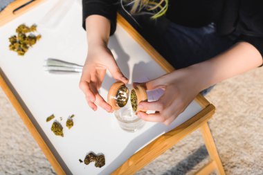 top view of girl putting weed from marijuana grinder in glass on wooden tray clipart