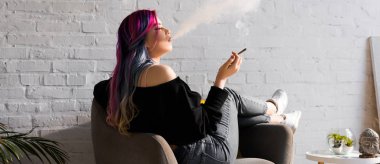 panoramic shot of hipster girl with colorful hair sitting in armchair and blowing smoke clipart