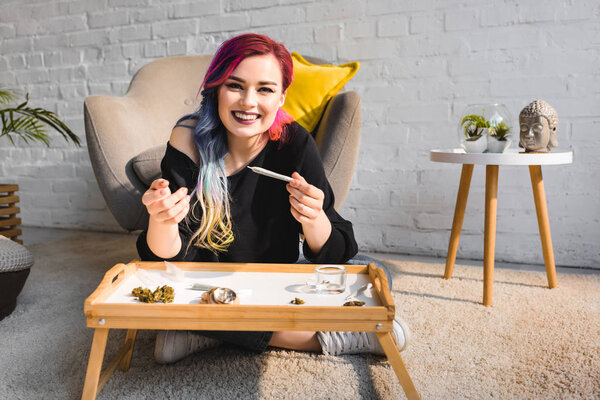 attractive hipster girl sitting on floor behind table, rolling joint and smiling at camera 