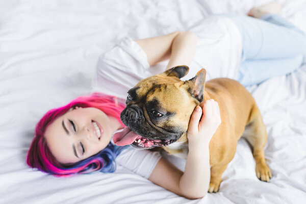 selective focus of french bulldog and happy girl with colorful hair laying with in bed 