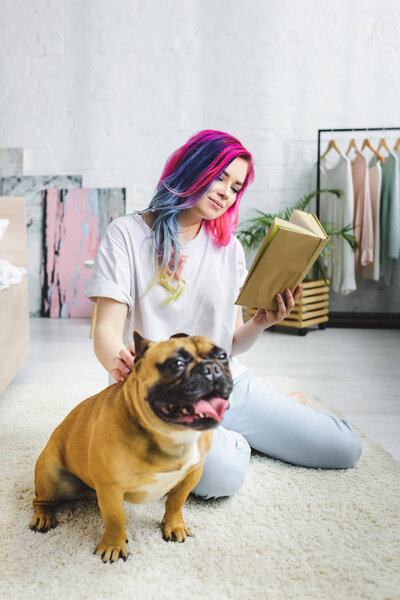selective focus of beautiful girl with colorful hair reading book and sitting on floor near French bulldog