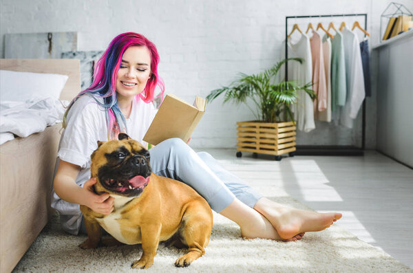  beautiful girl with colorful hair holding book, smiling and sitting on floor near French bulldog