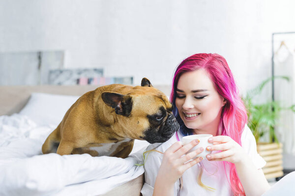 happy girl with colorful hair holding cup with coffee and cute bulldog sitting on bed and sniffing coffee