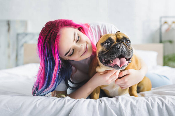 girl with colorful hair petting and looking at cute french bulldog while laying in bed