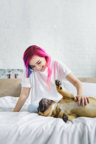 girl with colorful hair petting and looking at cute french bulldog, which laying on back in bed